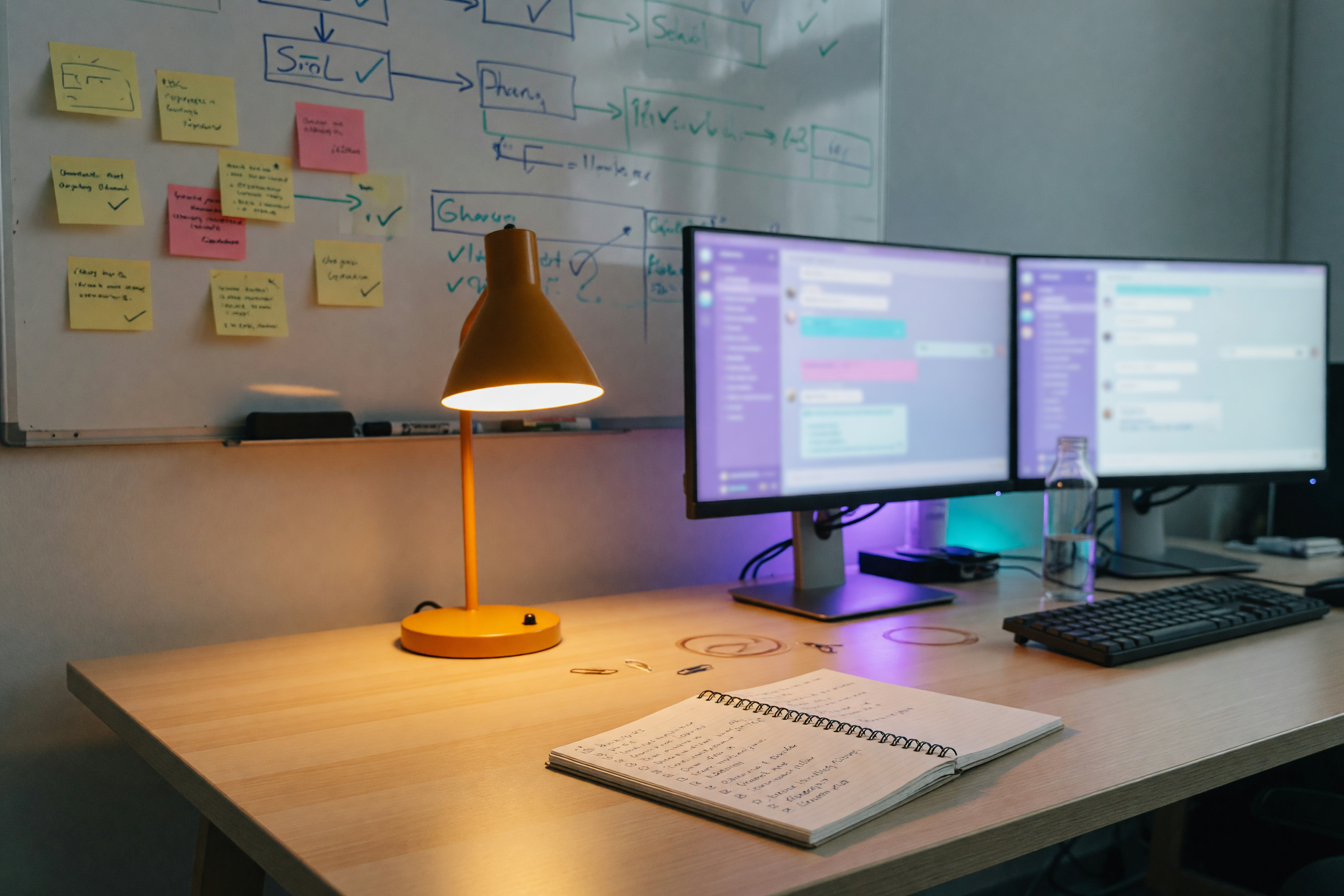 Espacio de trabajo de un equipo reducido con un monitor que muestra canales de Discord e hilos de chat activos, rodeado de notas adhesivas, una pizarra de proyecto y un cuaderno bajo una cálida luz ámbar de oficina