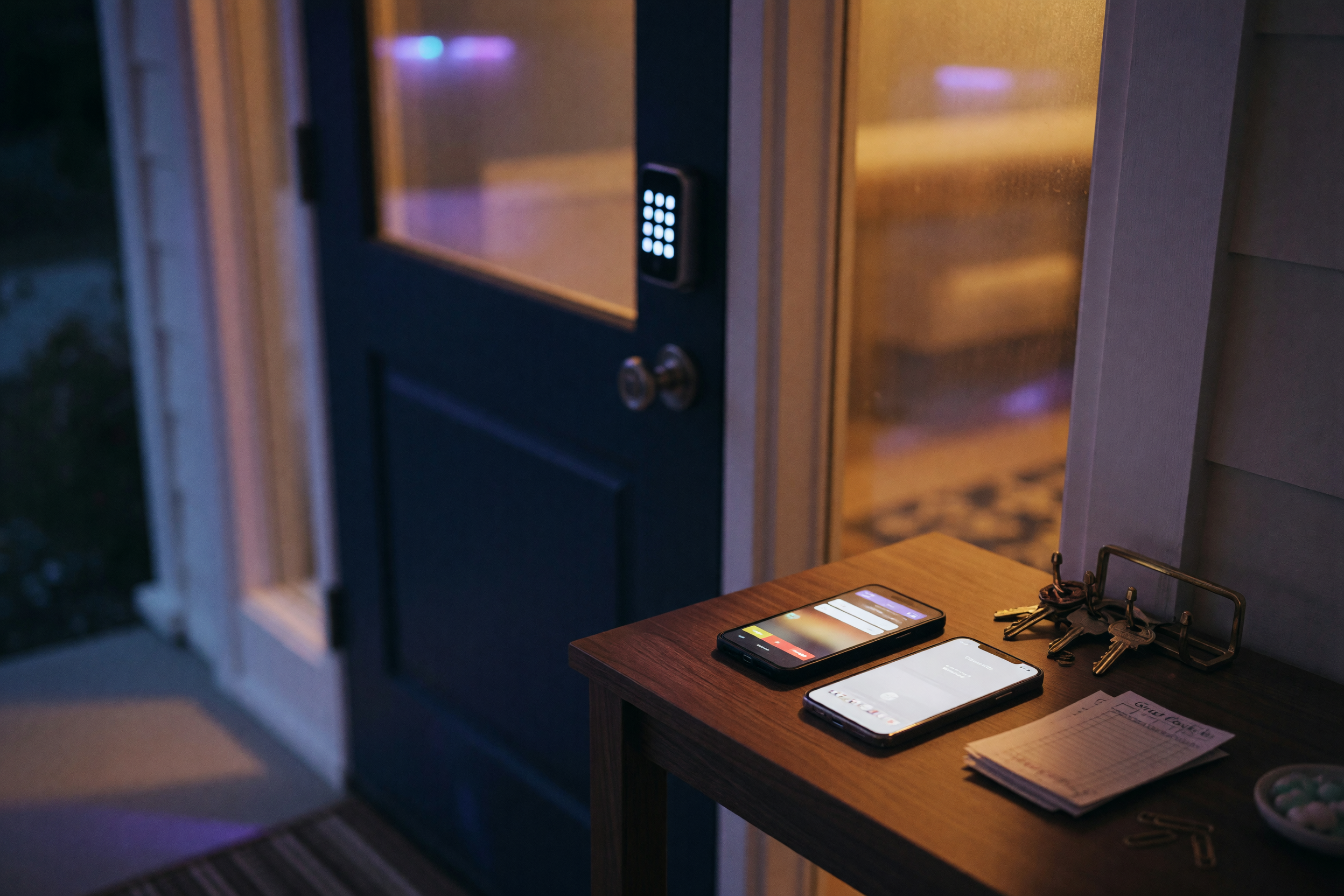 Vacation rental entryway at night with a glowing smart-lock keypad, a ringing phone on a wooden console table, warm amber interior light through frosted glass doors, soft violet ambient glow from outside